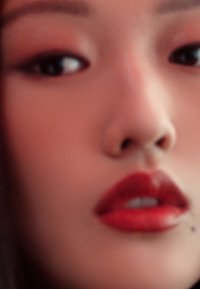 Gros plan sur un visage avec une peau lisse, des lèvres rouges pulpeuses et des yeux sombres. L'arrière-plan est flou, mettant en valeur les traits du visage et le maquillage.