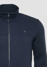 Marine blauwe ritsjack met hoge kraag, gemaakt van gladde stof. Voorzien van een metalen rits en een klein logo op de voorkant.