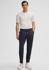 Polo shirt bianco a maniche corte, pantaloni blu slim-fit e sneaker bianche. I pantaloni presentano un leggero risvolto alla caviglia.