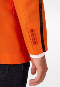 Męska dłoń obok rękawa pomarańczowego żakietu z czterema guzikami i czarnym paskiem, noszonego na białej koszuli i ciemnych dżinsach.