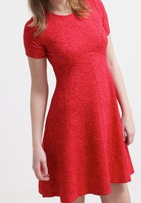 Röd kortärmad klänning med blommönster, figurnära upptill och utsvängd kjol. Slätt tyg med ett subtilt mönster genom hela plagget.