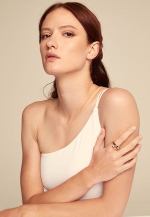 Joven con cabello rojo que lleva un top blanco de un solo hombro, sosteniendo su brazo izquierdo, adornado con un anillo grueso de oro.