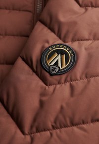 Brązowa puchowa kurtka z teksturowaną tkaniną, ukośnym pikowaniem oraz czarną okrągłą naszywką z logo „Superdry” z złotymi i białymi detalami.