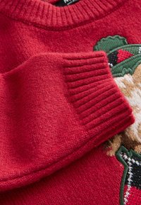 Pull en tricot rouge avec des poignets côtelés et un motif texturé agrémenté de touches vertes et crème. Détail en gros plan du tissu et du design.