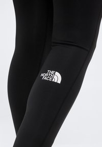 Κοντινό πλάνο μαύρων κολάν με λευκό λογότυπο The North Face στο μηρό, σε απλό ανοιχτό φόντο.