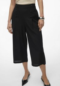Pantalons noirs raccourcis avec un détail de double bouton à la taille, des plis et une texture lisse. Associés à des chaussures noires pointues.