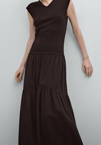 Musta mekko, jossa on istuva yläosa ja hihattomat hihat, siirtyen liikkuvaan kerrokselliseen hameeseen. Valmistettu pehmeästä kankaasta, jossa on sileä pinta.