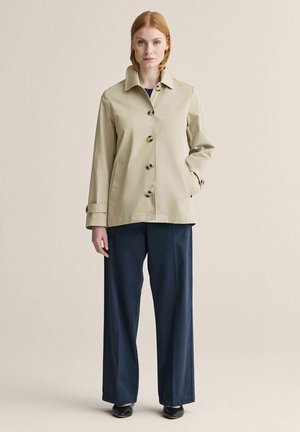 Kvinde med rødt hår iført en beige jakke med knapper over marineblå vidde bukser og sorte sko, stående med den ene hånd i lommen.