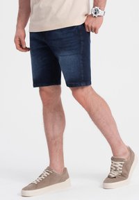 Shorts en denim bleu foncé avec une texture lisse, présentant une coupe standard et un ourlet retroussé, associés à des baskets en daim beige.