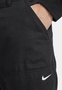 Musta Nike-housut, joissa on sivutasku ja valkoinen logo. Materiaali on sileä, jossa on hienovarainen textura, ja näkyvissä ovat saumat ja taskun yksityiskohdat.