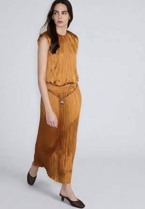 Donna che indossa un vestito senza maniche plissettato color marrone dorato con una cintura a catena decorativa, che cammina davanti a uno sfondo grigio chiaro uniforme.