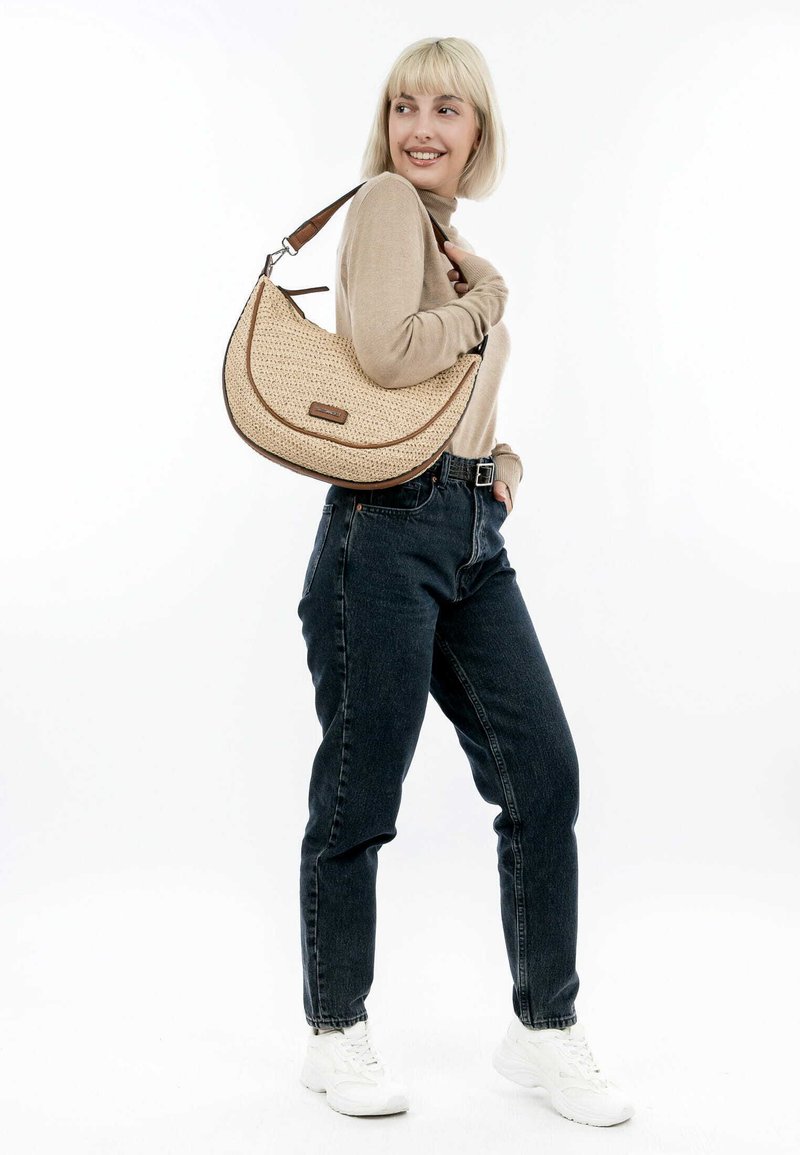 Sac à épaule tissé beige avec une forme courbée, garniture marron et une poignée supérieure. Le modèle l'associe avec un jean foncé et des baskets blanches.