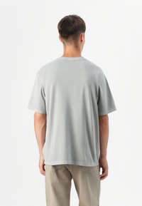 En mand står med ryggen til kameraet, iført en grå t-shirt og lyse bukser, foran en ensfarvet hvid baggrund.