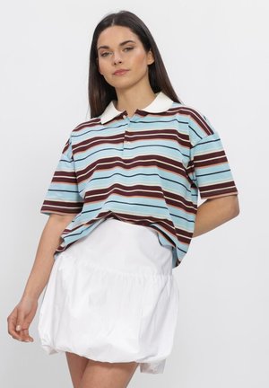 Vrouw met lang donker haar, gekleed in een blauw en bruin gestreepte poloshirt en een witte pofrok, staand voor een effen achtergrond.
