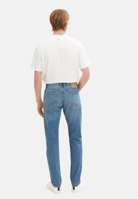 Jeans din denim albastru, cu croială dreaptă, cinci buzunare și o eticheta din piele maro pe talie, purtate cu un tricou alb simplu.