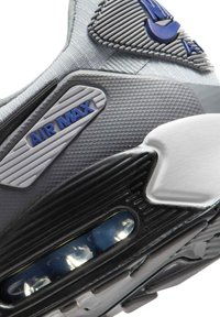 Primer plano de una zapatilla Nike Air Max en gris y negro que muestra el logo "AIR MAX", material texturizado y la amortiguación de aire visible en la suela.