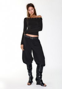 Schwarzes Off-Shoulder-Oberteil mit langen Ärmeln, kombiniert mit hoch taillierten schwarzen Hosen und glänzenden kniehohen Keilstiefeln. Auffällige silberne Beschläge am Gürtel.