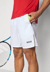 Un short de sport blanc avec un logo noir contrastant, une poche latérale qui contient une balle de tennis, et un design épuré et léger adapté aux activités sportives.