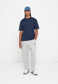 Osoba koja nosi plavu kapu, tamnoplavu majicu s kratkim rukavima, svijetlosive trenirke s natpisom i bijele tenisice s crnim prugama, stoji naslonjena na bijelu pozadinu.