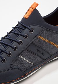 Mörkblå Rieker-sneakers med tyg- och läderdetaljer, orangea sömmar, mörkblå snören och en orange dragflik på tungan.