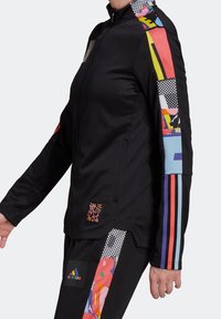 Black athletic jacket with zip, featuring colorful patterned sleeves and accents. Material appears smooth and stretchy. Notable logo on pocket.