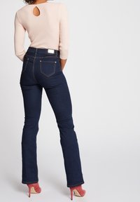Vaqueros acampanados de color azul oscuro con una cintura alta y dos bolsillos traseros. La blusa de canalé rosa claro tiene un pequeño detalle de ojal en la parte posterior.