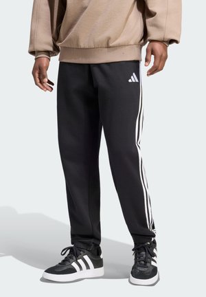 Osoba nosząca czarne spodnie dresowe Adidas z białymi paskami i logo, w połączeniu z czarno-białymi butami Adidas i beżową bluzą.
