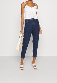 Bílý květinový crop top s nastavitelnými ramínky, tmavě modré džíny s vysokým pasem, bílé sandály na klínku a krémová kabelka. Hladké textury a čisté linie.