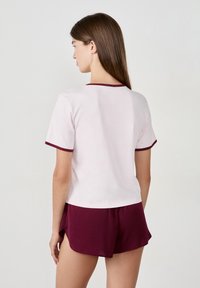 Donna vista di spalle che indossa una maglietta rosa chiaro a maniche corte con bordi scuri e pantaloni corti borgogna scuri su uno sfondo semplice.