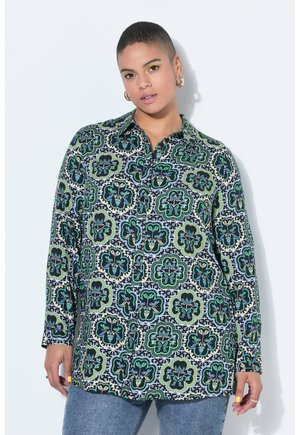 Person med kort hår iført en langærmet skjorte med grønne og blå blomsterprint og blå jeans.