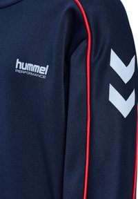 Marineblå sportjakke med røde kantbånd, hvidt "hummel PERFORMANCE" logo på brystet og to hvide vinkler på ærmet.