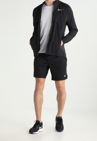 Czarna, lekka kurtka Nike z zapięciem na zamek, w połączeniu z czarnymi szortami i czarnymi butami sportowymi. Białe akcenty logo Nike na kurtce i szortach.
