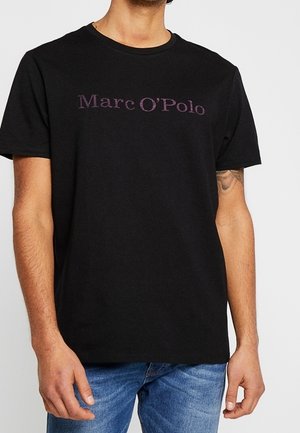 T-shirt en coton noir arborant le logo "Marc O'Polo" en violet sur la poitrine, à manches courtes et encolure ras du cou, porté avec un jean bleu.