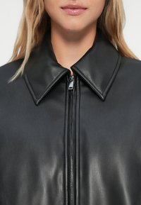 Nahaufnahme einer Frau in einer schwarzen Lederjacke mit weitem Kragen und vorderem Reißverschluss, Lippen sichtbar, aber Augen außerhalb des Rahmens.