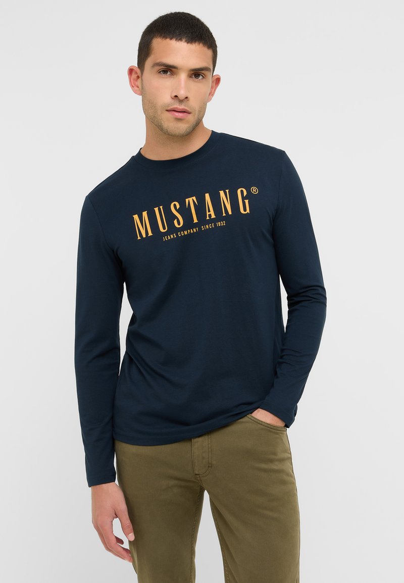 Hosszú ujjú, sötétkék pamut ing, amelyen egy nagy narancssárga "MUSTANG" logó található a mellrészen, valamint kerek nyakkivágás.