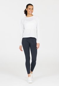 Vit långärmad topp med rund halsringning, kombinerad med åtsittande marinblå leggings, båda tillverkade av elastiskt tyg. Modellen har på sig vita sneakers.