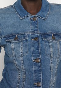Světle modrá džínová bunda s klasickým límcem, předním zapínáním na šest knoflíků a dvěma náprsními kapsami se mosaznými doplňky a viditelnými švy.