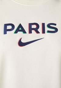 Tejido blanco con la palabra bordada "PARIS" en letras marinas contorneadas en azul, verde y rojo, encima de un logotipo de swoosh de Nike en color marino.