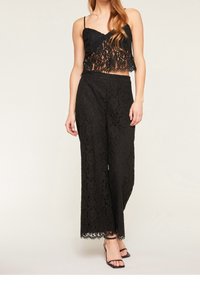 Svart spets crop top med tunna axelband över matchande svarta spets vida byxor. Båda har intrikata blommönster och vågiga kanter.