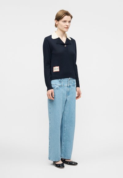 Μια γυναίκα στέκεται φορώντας μπλε σκούρο πουλόβερ με λευκή γιακά, ανοιχτό μπλε τζιν παντελόνι με φαρδιά μπατζάκια και μαύρα παπούτσια μπαλαρίνας με ασημένιες τρουκς, πάνω σε απλό φόντο.