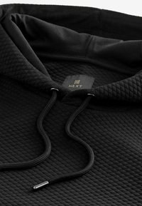 Zwarte gewatteerde hoodie met een trekkoordcapuchon, textuurstof en een label met het "NEXT"-logo in goud. Accenten omvatten zilveren aglets.
