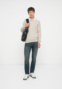 Kremfarget strikkegenser med ribbet tekstur, blå jeans og hvit skjorte-krage; holder en svart blank skulderveske, iført grå og hvite sneakers.