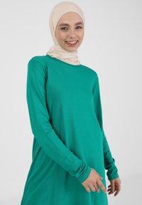 Robe verte à manches longues avec une coupe décontractée, fabriquée à partir d'un tissu doux. Parmi ses caractéristiques notables, on trouve des poignets avec ouverture pour le pouce et un col rond.
