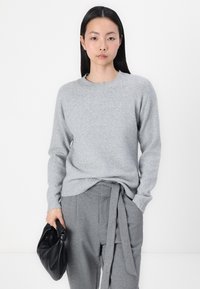 Grauer Pullover mit Rundhalsausschnitt und gerippten Bündchen, kombiniert mit grauen Hosen mit gebundenem Bund und einer schwarzen Handtasche.