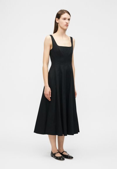 Γυναίκα που στέκεται φορώντας μαύρο midi φόρεμα χωρίς μανίκια με τετράγωνη λαιμόκοψη και μαύρα παπούτσια Mary Jane με ασημένιες τρύπες.