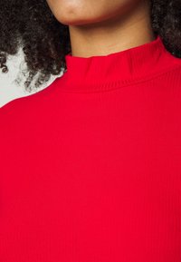 Haut rouge côtelé à col montant avec un tissu texturé. Le col présente un bord plissé, créant une silhouette structurée.