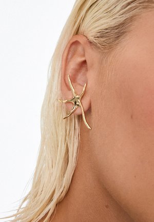 Pendiente con forma de estrella de mar dorada, llevado en la oreja izquierda de una persona con cabello rubio y mojado, sobre un fondo blanco liso.