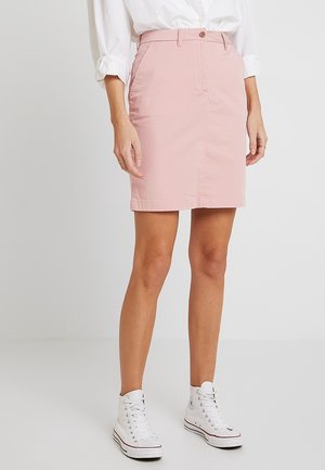 Femme portant une jupe rose jusqu'aux genoux, une chemise blanche boutonnée avec manches retroussées et des baskets montantes blanches, debout devant un fond uni.