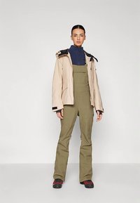 Beige wasserdichte Jacke mit schwarzen Akzenten über einer blauen Zwischenschicht und olivgrünen Latzhosen, beide mit Taschen und lockerem Sitz.