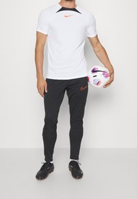 Biała koszulka Nike z krótkim rękawem i czarnymi akcentami, w połączeniu z czarnymi spodniami Nike z pomarańczowym logo; model trzyma piłkę nożną.